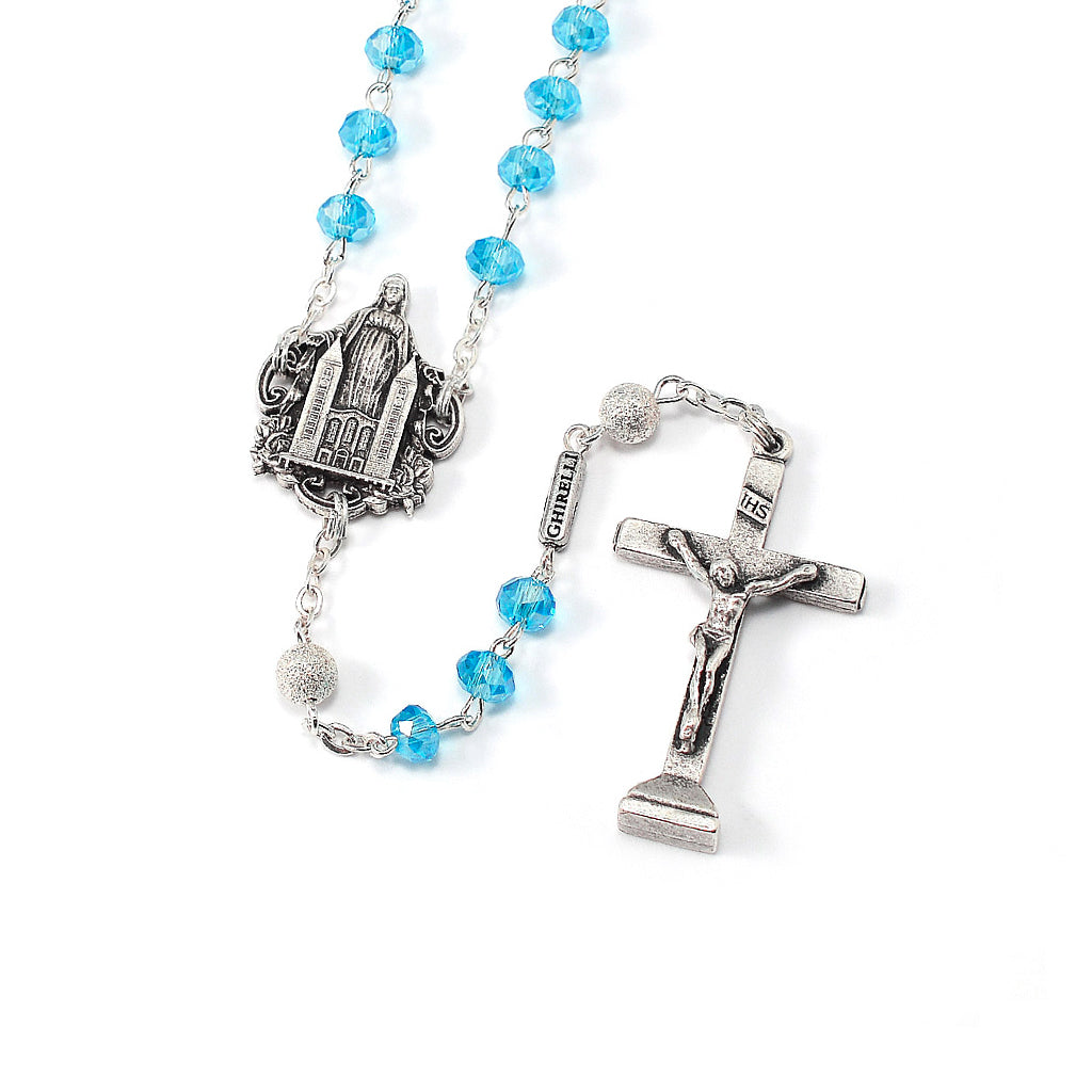 A rosary with aqua blue crystal beads and silver color metal accents, featuring a crucifix and a centerpiece with Mary, Queen of Peace.