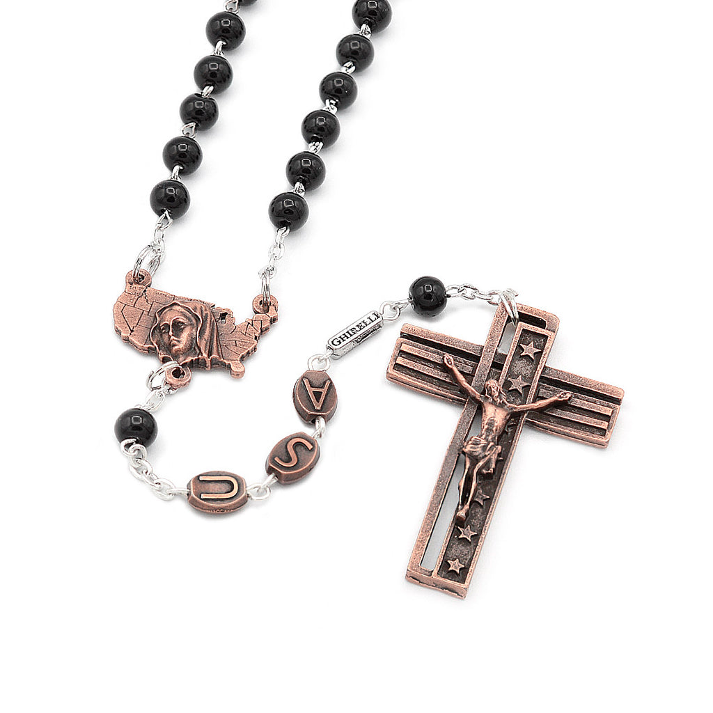 A rosary with black glass beads and antique copper finish, featuring a crucifix with an inscription and a centerpiece with an image of the Immaculate Conception.